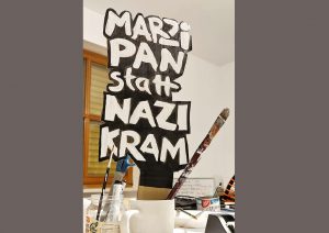 Das Bild zeigt ein handgemaltes Schild, das aus Pappe oder Karton besteht und mit schwarzer und weißer Farbe bemalt ist. Die Aufschrift „MARZIPAN statt NAZI KRAM“ ist in großen, ungleichmäßigen Buchstaben geschrieben, die einen handgemachten und etwas improvisierten Eindruck vermitteln. Die weiße Farbe auf dem schwarzen Hintergrund sorgt für einen starken Kontrast und macht die Botschaft deutlich lesbar. Die Stimmung ist klar politisch; das Bild ist eine Aussage gegen den Nazismus und für Toleranz und Frieden. 