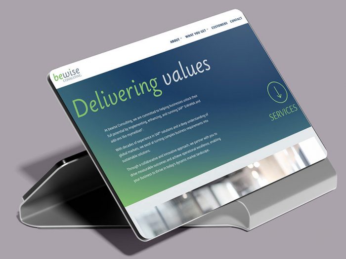 Das Bild zeigt einen Tablet-Ständer mit einem iPad darauf, auf dem die Webseite von bewise Consulting angezeigt wird. Die Webseite selbst präsentiert sich modern und übersichtlich und wirbt mit den Kernkompetenzen des Unternehmens: Beratung und Implementierung von SAP S/4HANA und Add-ons. Der Fokus liegt auf dem Wertversprechen ("Delivering Values") und dem partnerschaftlichen Ansatz zur Erreichung messbarer Erfolge für Kunden.